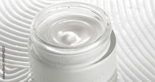 Open jar with cosmetic facial cream on a background of cream texture, close-up on a rotating table.