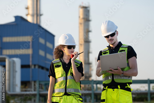 Two petroleum engineers in hardhat and safety vest reviewing blueprint and pointing at oil refinery industrial site during sunset for inspection teamwork planning discussion in energy industry.