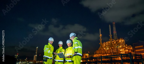 Petroleum engineers team wearing safety uniform and hardhat with laptop monitoring and discussing work at oil refinery site during night operation engineering teamwork in energy industry.