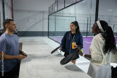 Diverse players standing at marble counter beside glass padel court holding rackets, cup, ball