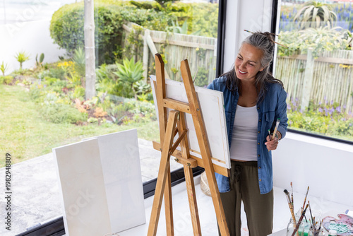 Senior woman wearing denim shirt, olive pants standing at easel painting with palette, copy space