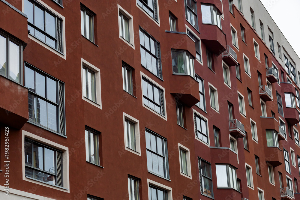 Fototapeta premium Red brick apartment building rises tall. Windows reflect sky and clouds. Balconies curve around each unit. Building shows urban residential life. Ideal for cityscape or housing themes