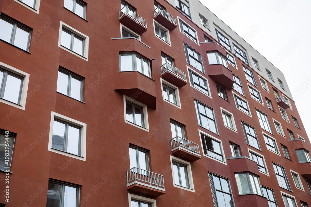 Obraz premium Red brick building with many windows and balconies. Balconies feature metal railings, some closed, others open. Windows reflect sky and clouds, adding depth. Architecture shows urban living