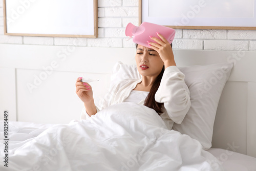 Young Asian woman with thermometer and hot water bottle warming her head on b...