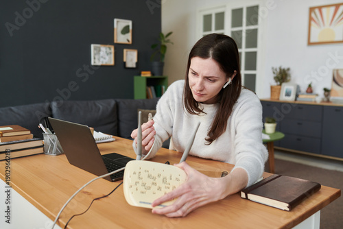 Photos Caucasian young adult woman connecting router cable beside laptop, troubleshooting home internet setup for remote work and digital access