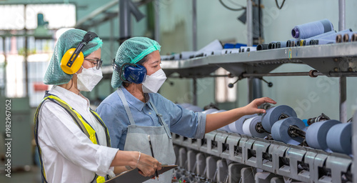 Textile inspection factory teamwork female supervisor in safety vest checking and discussing yarn production quality with woman worker in textile mill holding thread spool and clipboard.
