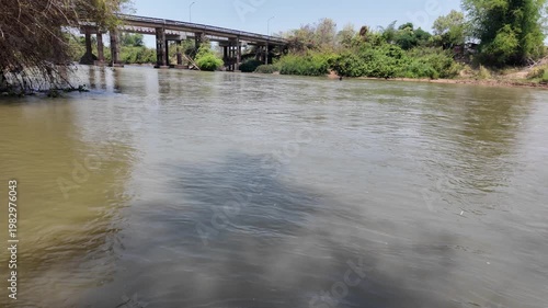 Serene river flows beneath bridge, with calm waters reflecting lush greenery and trees, creating tranquil outdoor landscape that embodies beauty of nature.