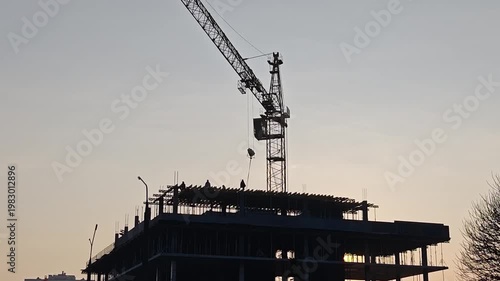 Silhouette of a construction site with a towering crane and workers on a building frame against a sunset sky, with the sun peeking through the structure.