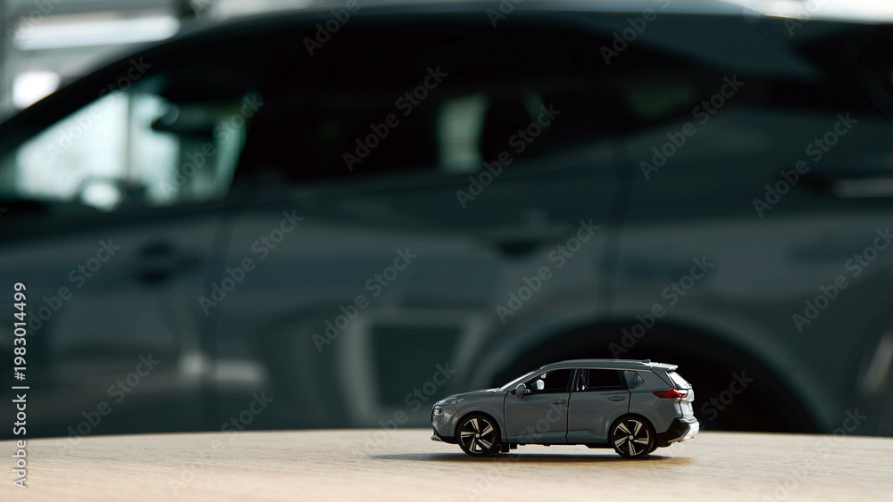 Fototapeta premium Miniature gray SUV model displayed on wooden surface with blurred full-sized gray SUV in the background, showcasing automotive design and scale comparison