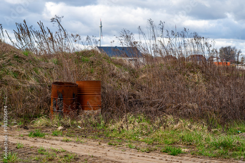 Konstfotografi Two old rusty metal barrels abandoned on a wasteland near a construction site, environmental pollution in rural area, residential houses and wind turbines in the background under cloudy sky