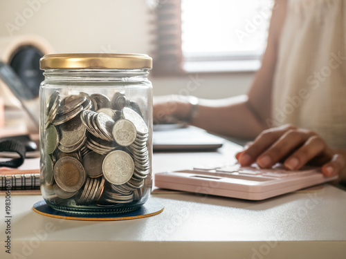 Glass jar filled with coins on desk with person using calculator. Saving money, financial planning, taxation, budgeting, personal finance, investment concept, home economy and wealth growth strategy.