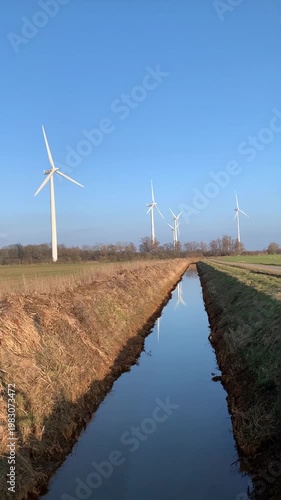 view of canal and field with rotating wind turbines against green meadow and blue sky early spring or late autumn