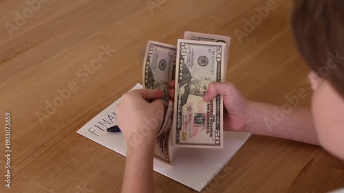 Child counting US dollar cash and writing finance notes on paper on beige wooden table. Early financial literacy, budgeting skills and money awareness concept with copy space indoors