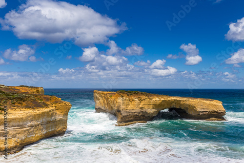 Surreal cliffs and rock formation London Bridge rise from the vibrant ocean, with waves crashing on sandy shores at Great Ocean Road, Victoria, Australia.