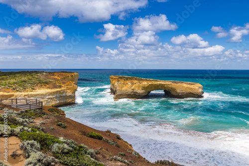 Surreal cliffs and rock formation London Bridge rise from the vibrant ocean, with waves crashing on sandy shores at Great Ocean Road, Victoria, Australia.