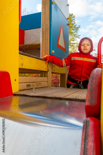 Child with red raincoat of firefighter clings to a rope in a playground during rain. High quality photo