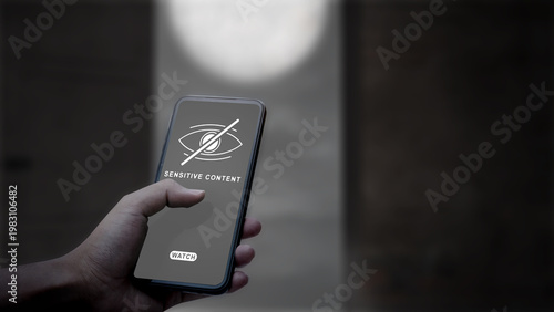 A person's hand holding a smartphone displaying a sensitive content warning message with a hidden eye icon, representing internet censorship and online safety