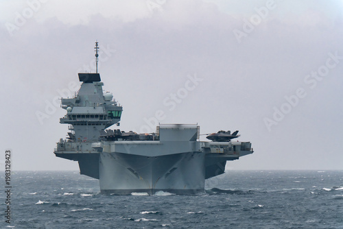 Royal Navy aircraft carrier HMS Prince of Wales sailing in Tokyo Bay, Japan, with F-35B fighter jets on deck, rough and cloudy weather.