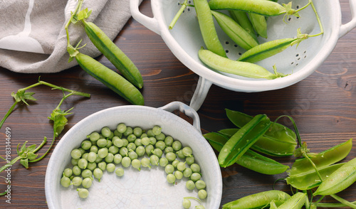 Fresh organic green peas being shelled in ceramic bowls on dark wooden table with copy space. High-angle shot features pea pods and individual peas highlighting seasonal harvest and healthy farm-to