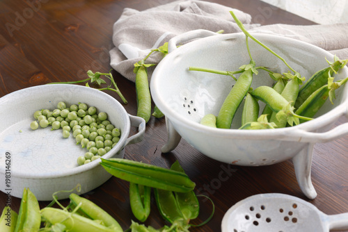 Fresh organic green peas being shelled in ceramic bowls on dark wooden table with copy space. High-angle shot features pea pods and individual peas highlighting seasonal harvest and healthy farm-to