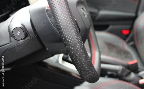 A steering column with a sports steering wheel against the background of blurred front seats with red details in the car interior
