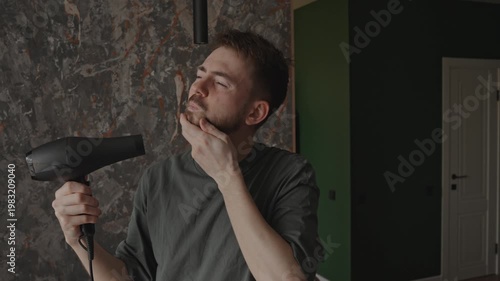 Man blowing hair with dryer, playful model vibe in modern interior, posing with confident smile and dramatic lighting, casual