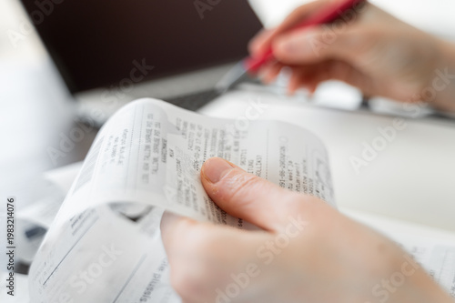 Hand holding receipt with another hand holding pen, more receipts, notebook and laptop visible in background, personal finance and accounting