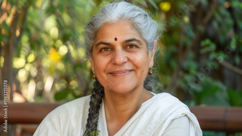 Smiling older woman with gray hair braided, wearing traditional white attire, sits outdoors with greenery in the background. Her gentle expression conveys warmth and calmness