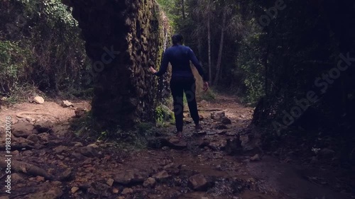 Mujer caminando con cuidado entre piedras y agua en un bosque frondoso con un acueducto