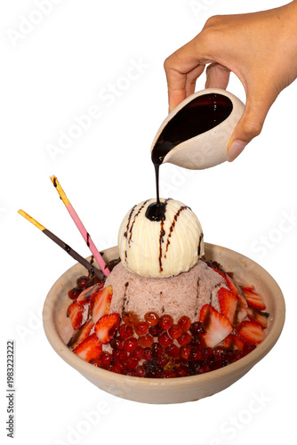 A hand pours dark chocolate syrup over a decadent bowl of chocolate shaved ice topped with ice cream and strawberries.