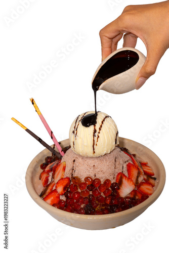 A hand pours dark chocolate syrup over a decadent bowl of chocolate shaved ice topped with ice cream and strawberries.
