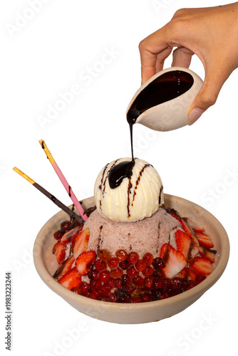 A hand pours dark chocolate syrup over a decadent bowl of chocolate shaved ice topped with ice cream and strawberries.