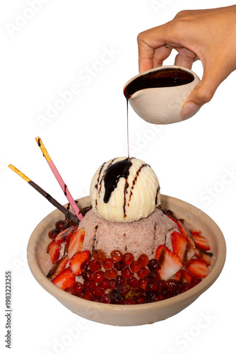A hand pours dark chocolate syrup over a decadent bowl of chocolate shaved ice topped with ice cream and strawberries.