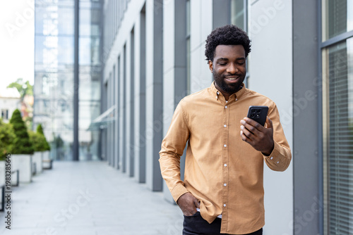 African person smiling while engaging with a mobile device outside a modern office building, representing communication, technology, and connection in a busy city setting