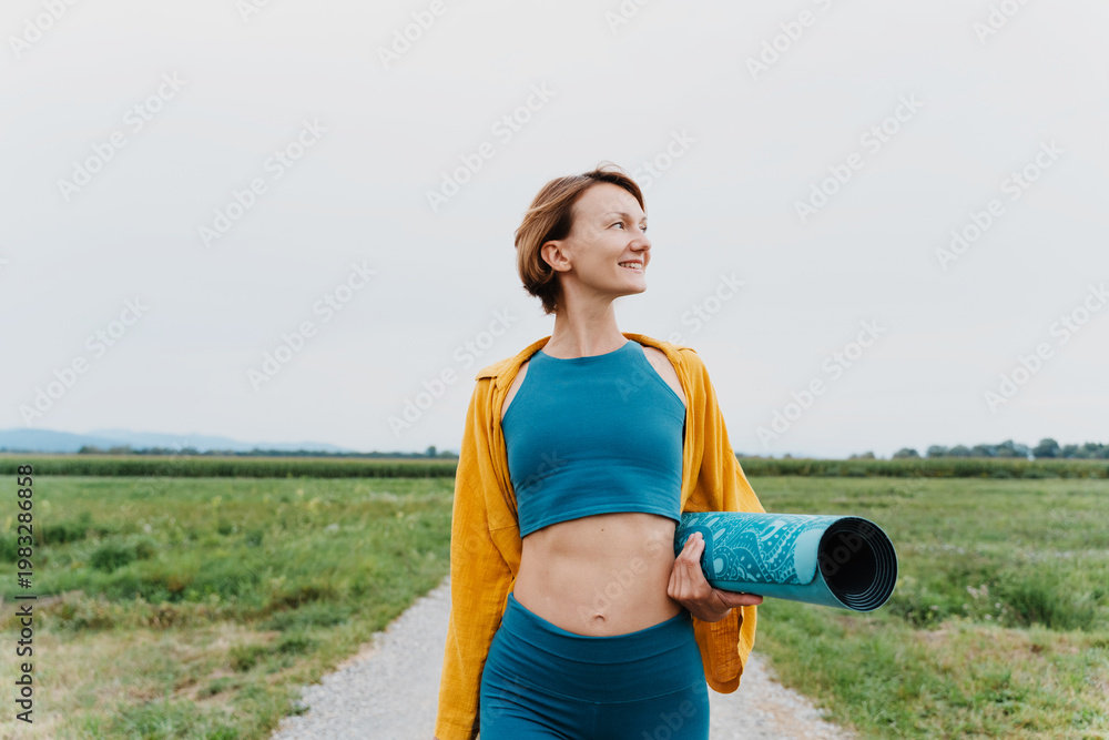 Obraz premium Beautiful fit woman holding yoga mat outdoors. Smiling female with exercise mat in green nature park