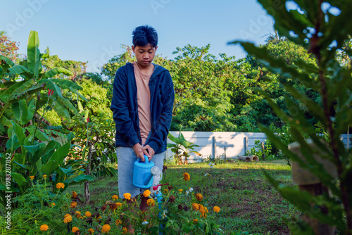 Asian Teenage Boy Standing in Garden Holding Blue Watering Can Among Marigold Flowers