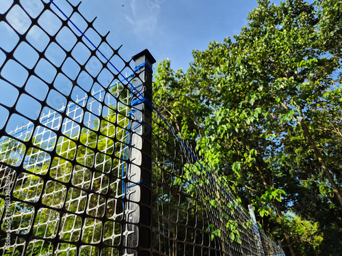 Obraz na plátně Low angle view of black mesh fence with green trees and blue sky background