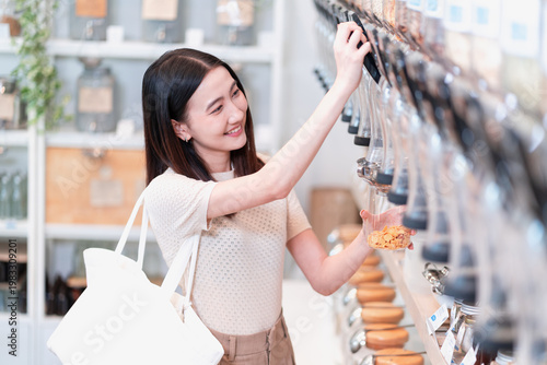 Foto Stylish young Asian woman with a white tote bag smiles as she reaches up to choose food from a wall of bulk dispensers in a zero waste shop