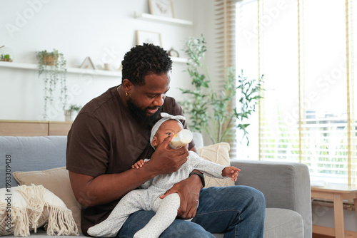 Foto Tender moment as a loving African American father holds and bottle-feeds his infant daughter while sitting on a cozy sofa in a bright living room