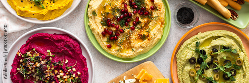 A vibrant spread showcases different types of hummus including classic, beetroot, broccoli, and turmeric. Accompanied by olives and breadsticks, it invites sharing and enjoyment, banner
