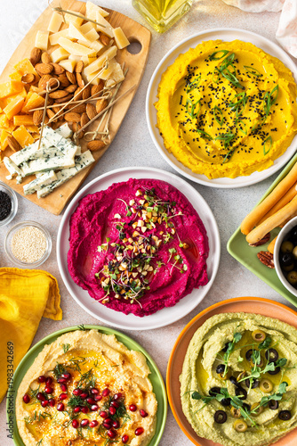 Colorful spread features beetroot, classic, broccoli, and turmeric hummus showcased on a charcuterie board with vibrant toppings, breadsticks, and assorted olives, perfect for gatherings