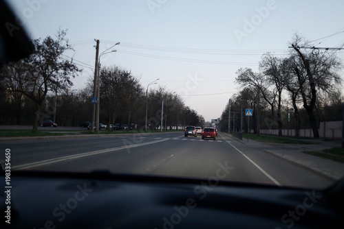 Forward view from inside a moving car along a quiet city road, soft evening light and distant vehicles creating depth, capturing routine travel and a calm, understated urban rhythm