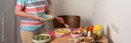 A 53-year-old woman is preparing dinner at home. She is setting the table while holding a pan with food. The setting is simple and warm, reflecting a typical evening meal time, banner