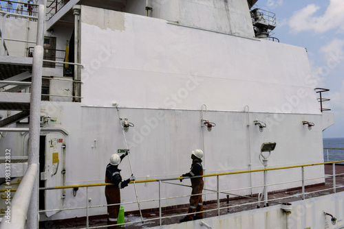 seafarers applying fresh paint on vessel superstructure using long rollers, showcasing routine maintenance, safety gear and teamwork at sea