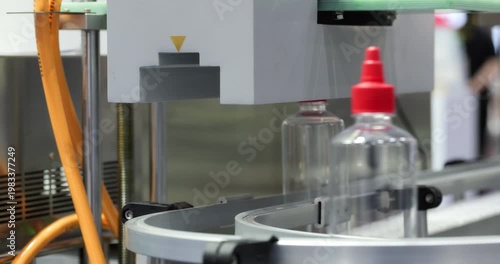 Plastic bottles with red caps move along conveyor under filling unit in sauce production line. Industrial food packaging process with automated control and modern equipment.