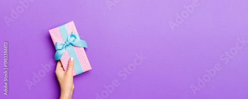Flat lay of woman hands holding gift wrapped and decorated with bow on purple background with copy space. Christmas and holiday concept