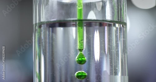 Close up of green liquid drops falling into clear solution inside glass container. Laboratory experiment showing mixing process, chemical reaction and scientific testing environment.