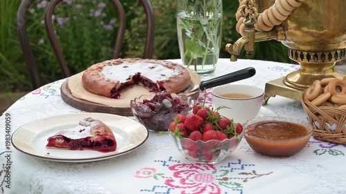 A rustic tea setting with golden samovar, fresh strawberries, jam, pie, and bagels arranged on an embroidered tablecloth outdoors.