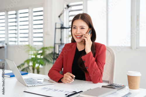 Happy joyful confident asian business lady, broker or officer, sitting at table in  office, working with laptop, talking to client or employees on smartphone, smiling