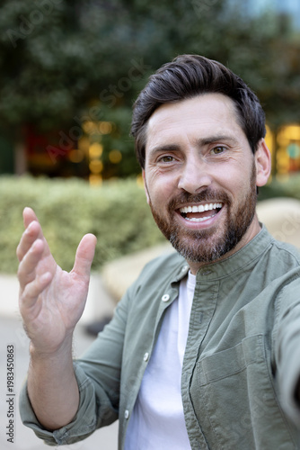 Cheerful man with a beard and dark hair smiles broadly and waves his hand while capturing a selfie outdoors, embodying happiness, welcome, and positivity